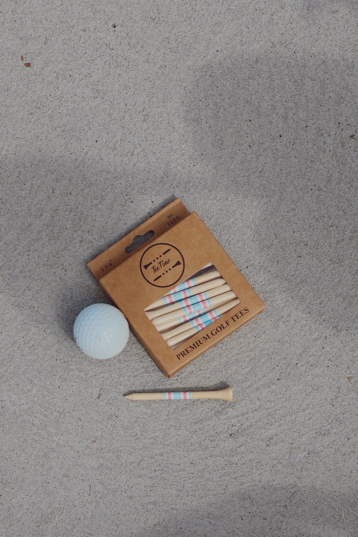 A Tee Time Golf box of pink and blue striped bamboo tees beside a golf ball and a loose tee on concrete.